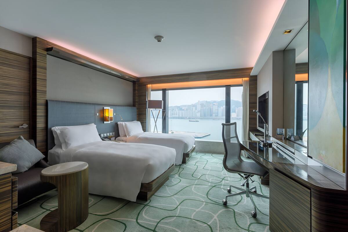 Une chambre d’hôtel avec deux lits, un bureau, une chaise, une télévision et une grande fenêtre donnant sur la ville.