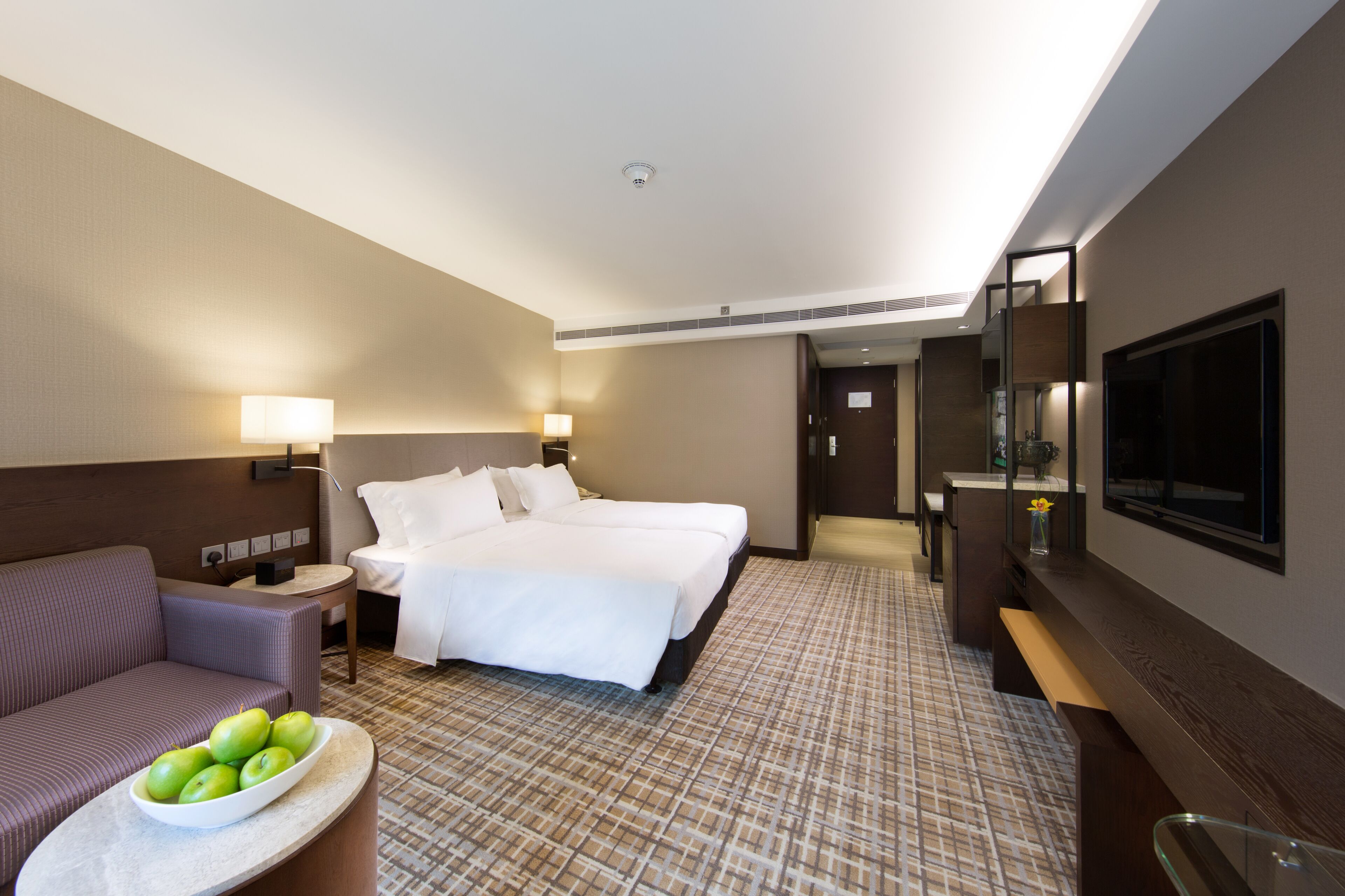 Habitación de hotel con cama, sofá, una mesita con manzanas, televisor y espejo.