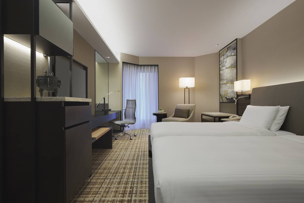 Une chambre d’hôtel avec deux lits, un bureau, une chaise, une lampe et un tableau au mur.