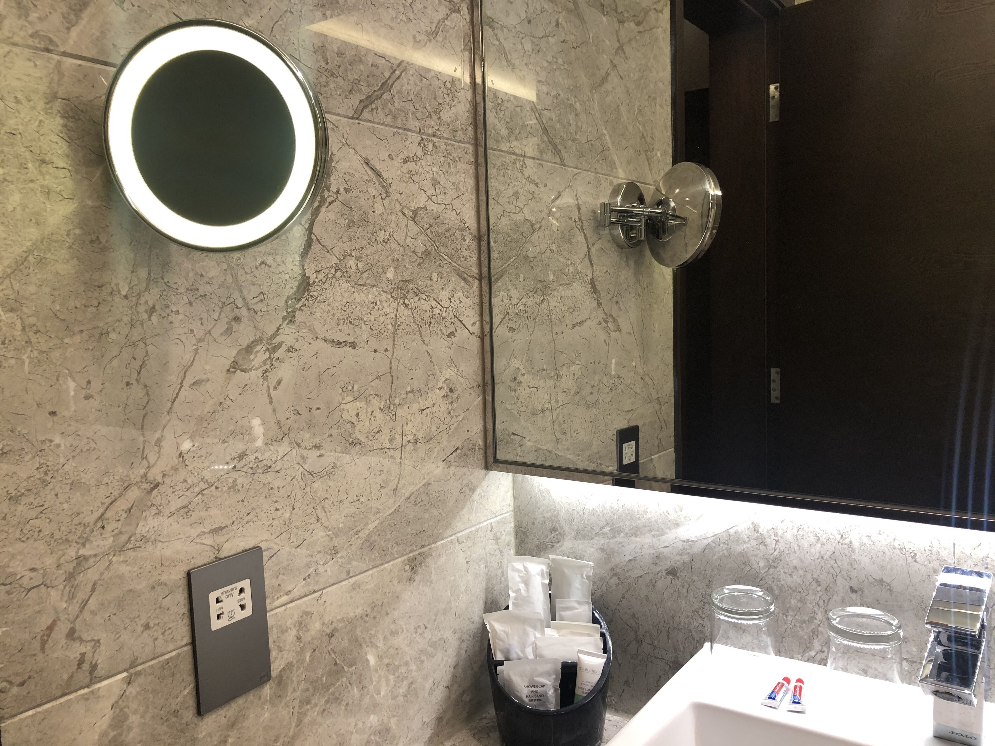 Un baño moderno con pared de mármol, un espejo redondo, ducha con puerta de vidrio y zona de lavamanos con artículos de aseo.
