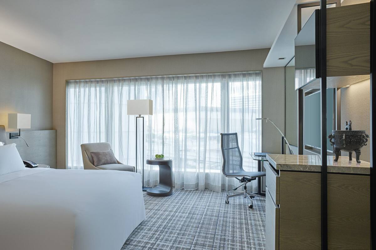 Une chambre d’hôtel moderne, dotée d’un grand lit, d’un bureau, d’une chaise, d’une petite table avec une lampe et d’une grande fenêtre ornée de rideaux légers.
