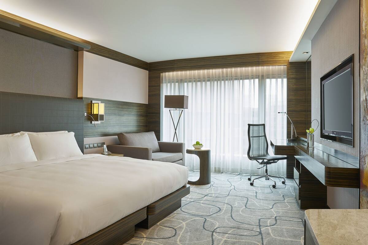 Une chambre d’hôtel moderne dotée d’un grand lit, d’un bureau, d’une chaise, d’une télévision et d’une grande fenêtre avec des rideaux.