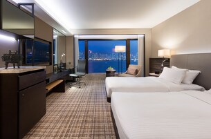 A hotel room with two beds, a desk, a chair, a TV, and a view of a cityscape at night.
