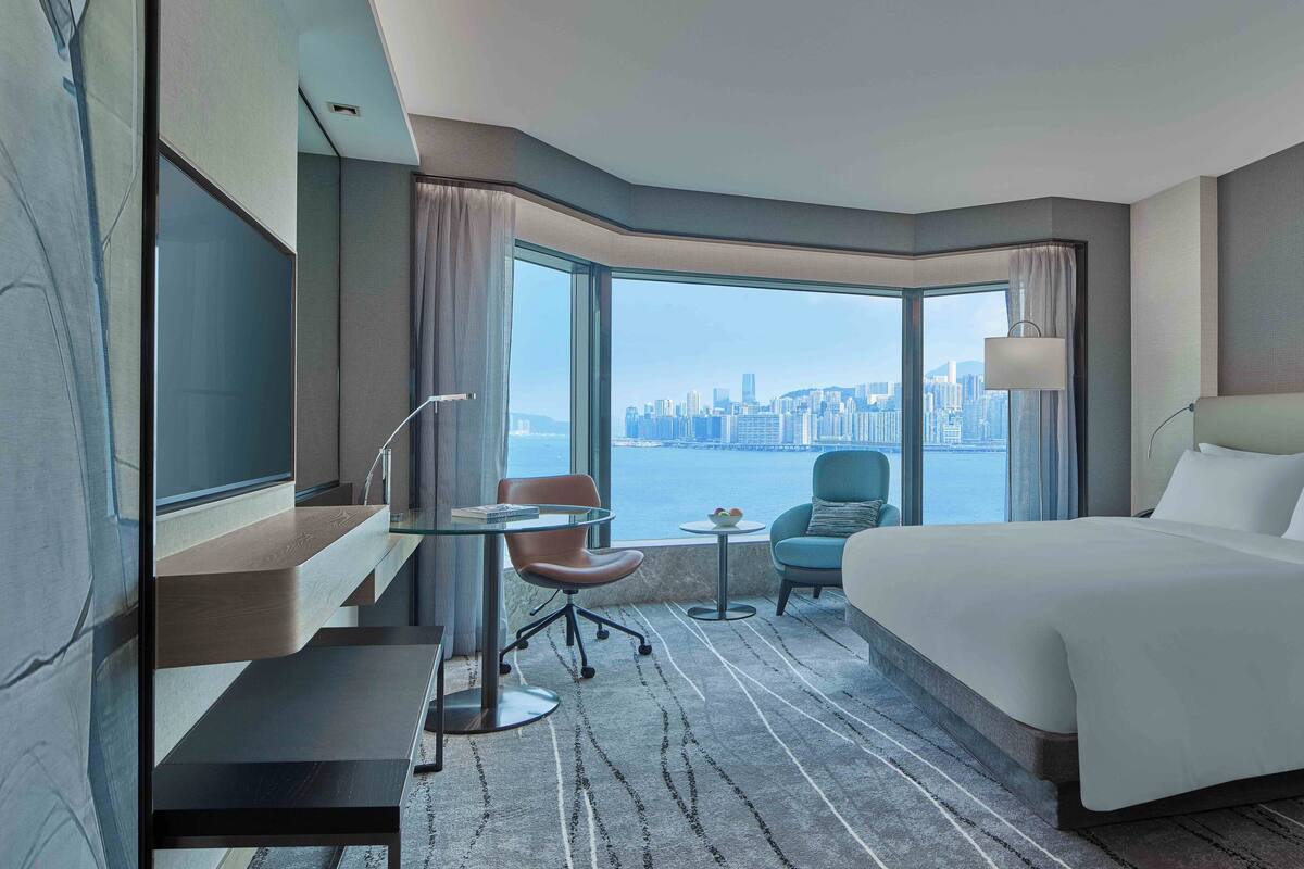 Une chambre d’hôtel avec un grand lit, un bureau avec une chaise, une petite table sur laquelle se trouve un saladier rempli de fruits, et une vue sur le panorama urbain à travers de grandes fenêtres.