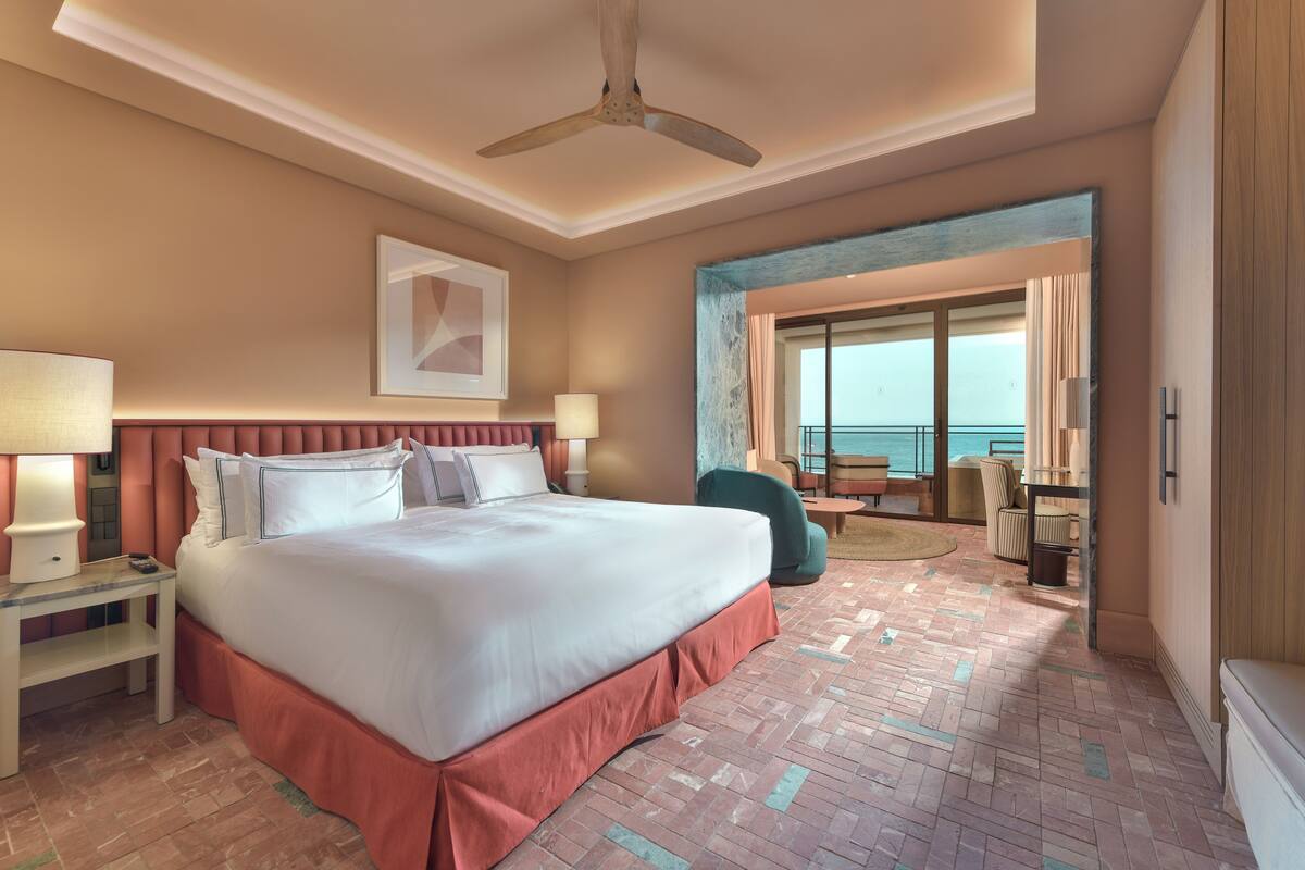 A hotel room with a large bed, a ceiling fan, a view of the ocean, and a balcony with outdoor seating.