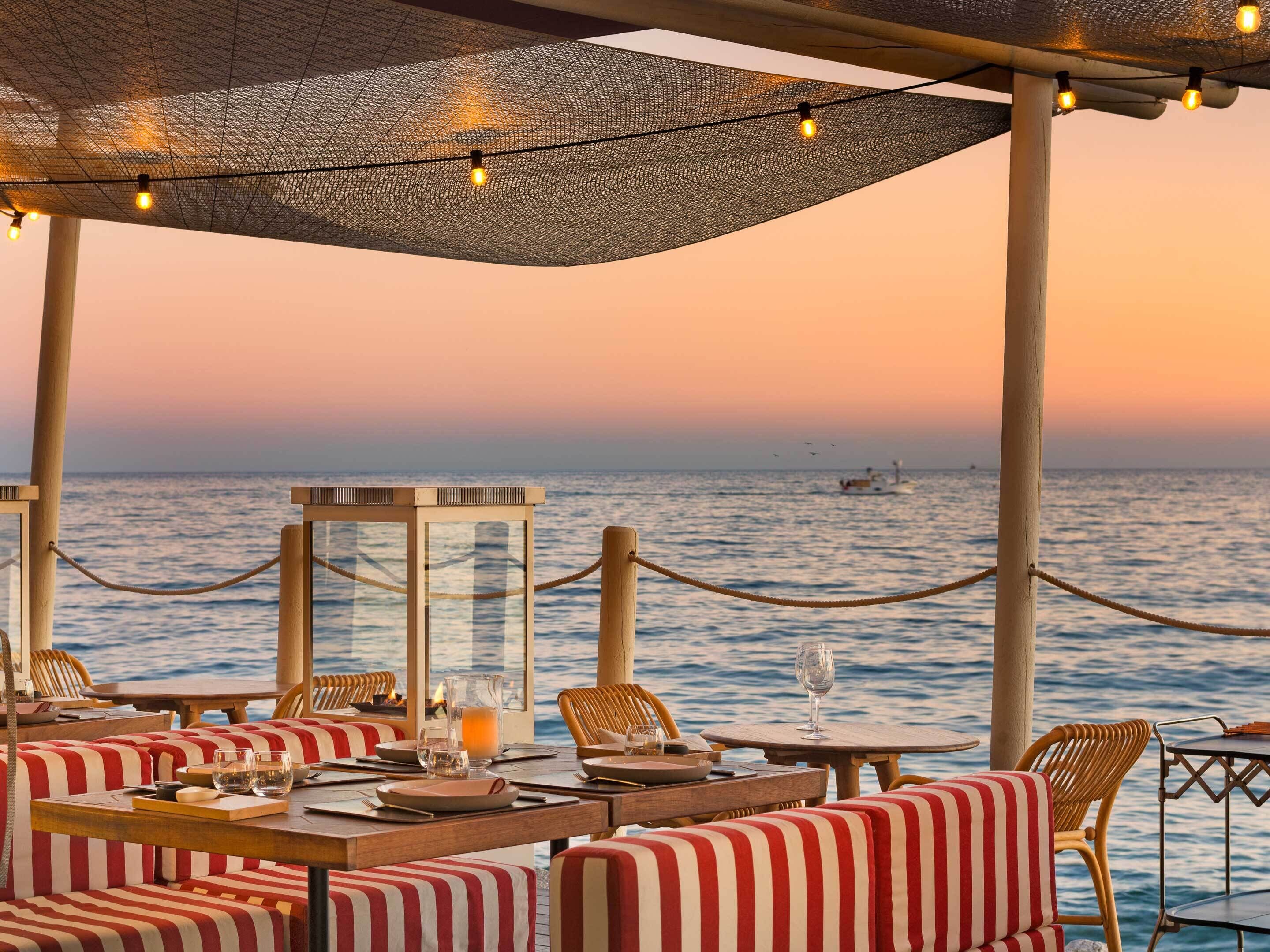 Un restaurante frente al mar con terraza, vista al océano y un atardecer.
