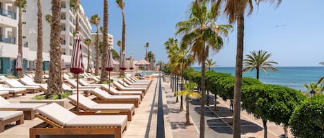 A beachfront hotel with rows of sun loungers and palm trees, a clear sky, and the ocean in the background.