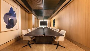 Uma sala de conferências com uma mesa comprida, cadeiras brancas e uma tela grande na frente.