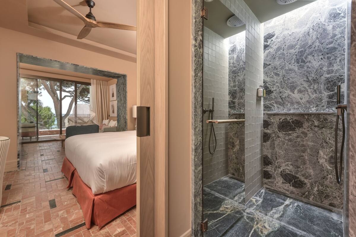 A bedroom with a large bed, a balcony with a view, and a shower with marble walls.