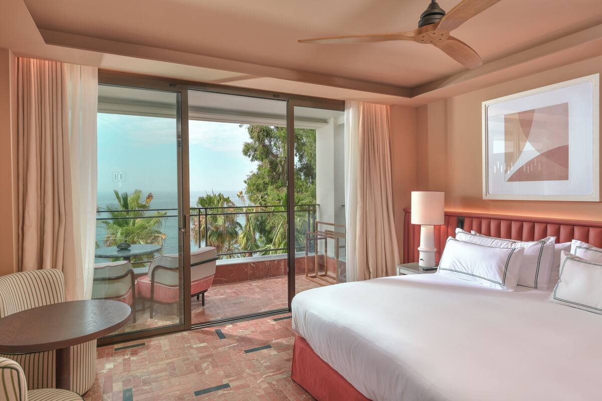 A hotel room with a bed, a balcony with a table and chairs, and a view of the ocean.