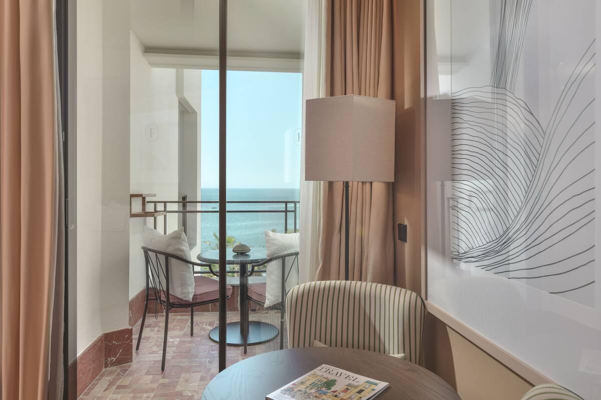 A balcony with a table and chairs, a view of the sea, and a large abstract painting on the wall.