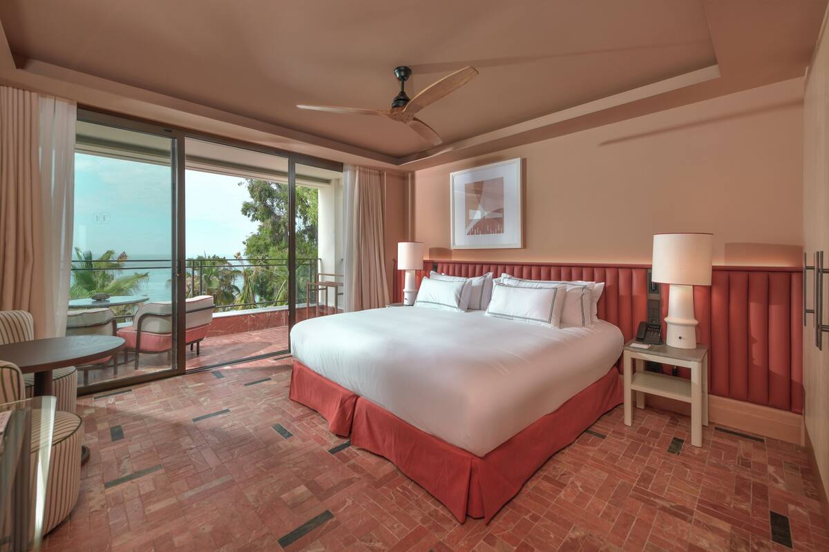 A spacious bedroom with a large bed, a ceiling fan, a bedside table with a lamp, and a view of the outdoors through a sliding glass door.
