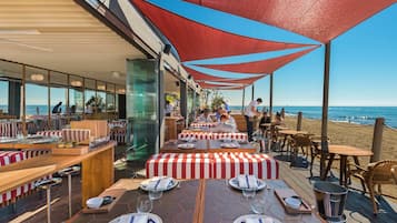 An outdoor dining area with tables set for two, red and white striped cushions, and a view of the beach and ocean.