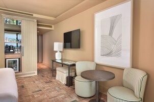 A hotel room with a bed, a small table with a lamp, a TV, a framed artwork, and a minibar.