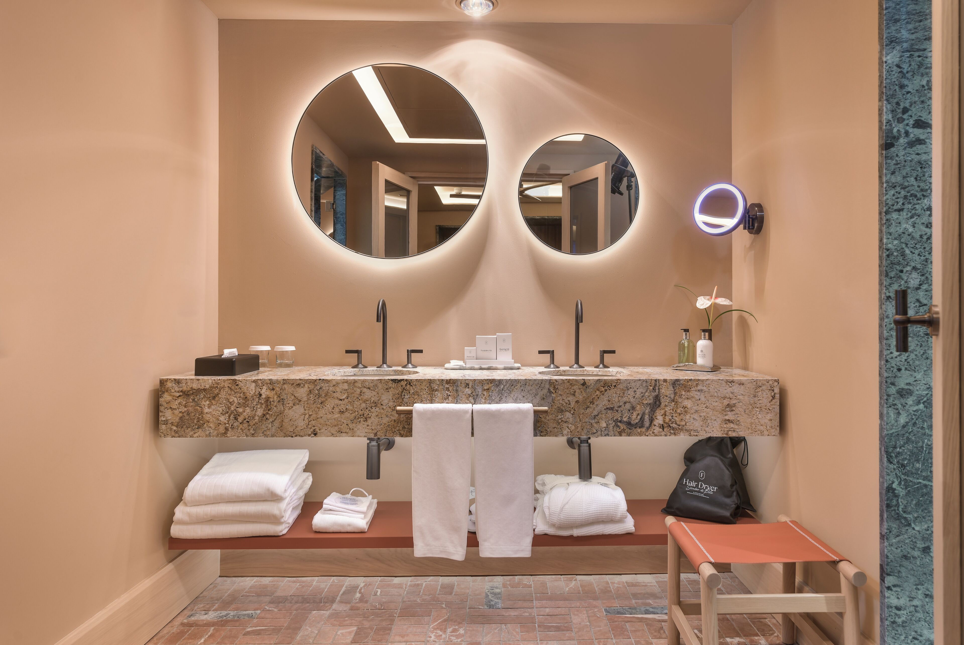 A bathroom with a double sink, two round mirrors, a vanity with a marble countertop, and a bench with towels.