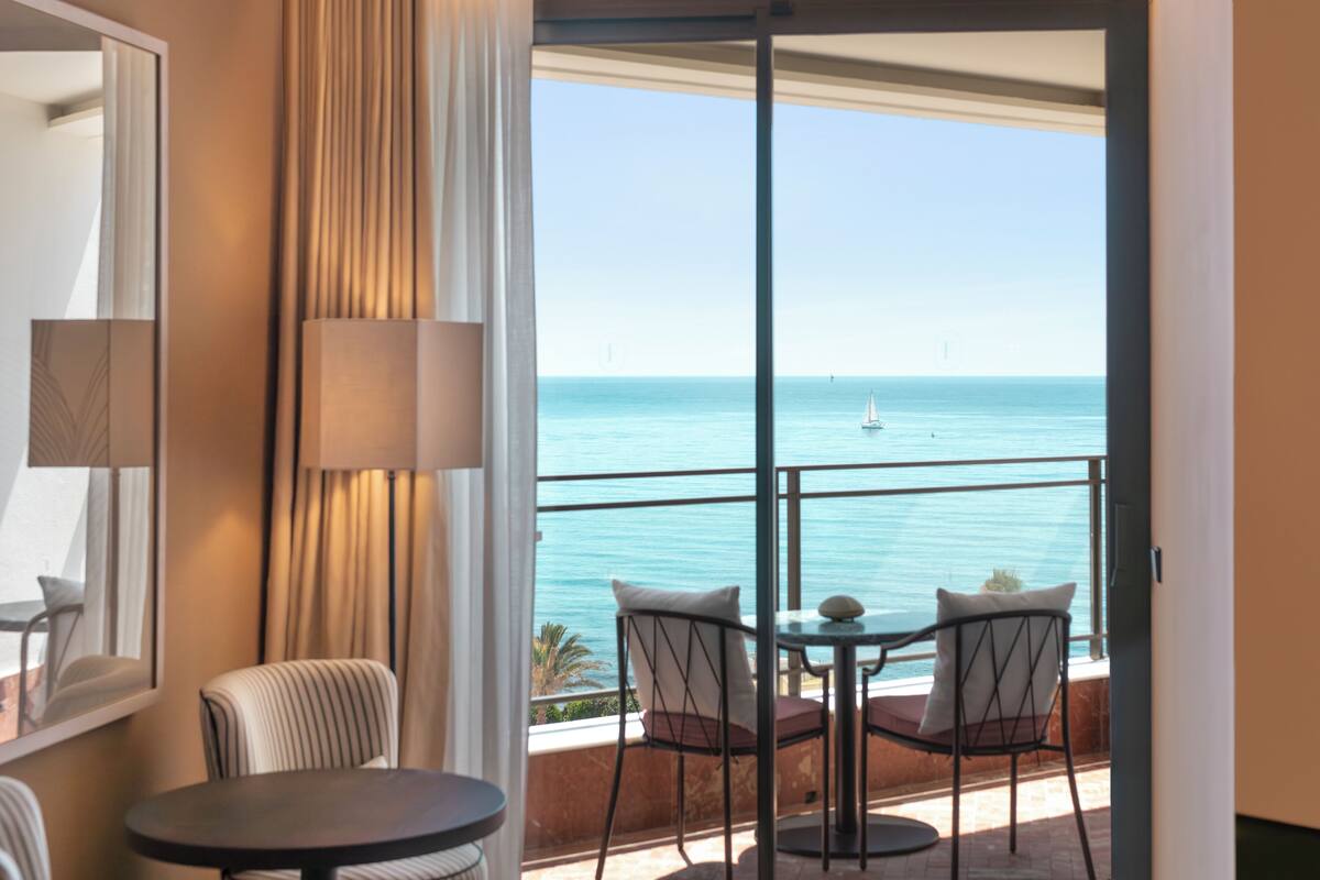 A balcony with a view of the sea, a table, two chairs, and a lamp.