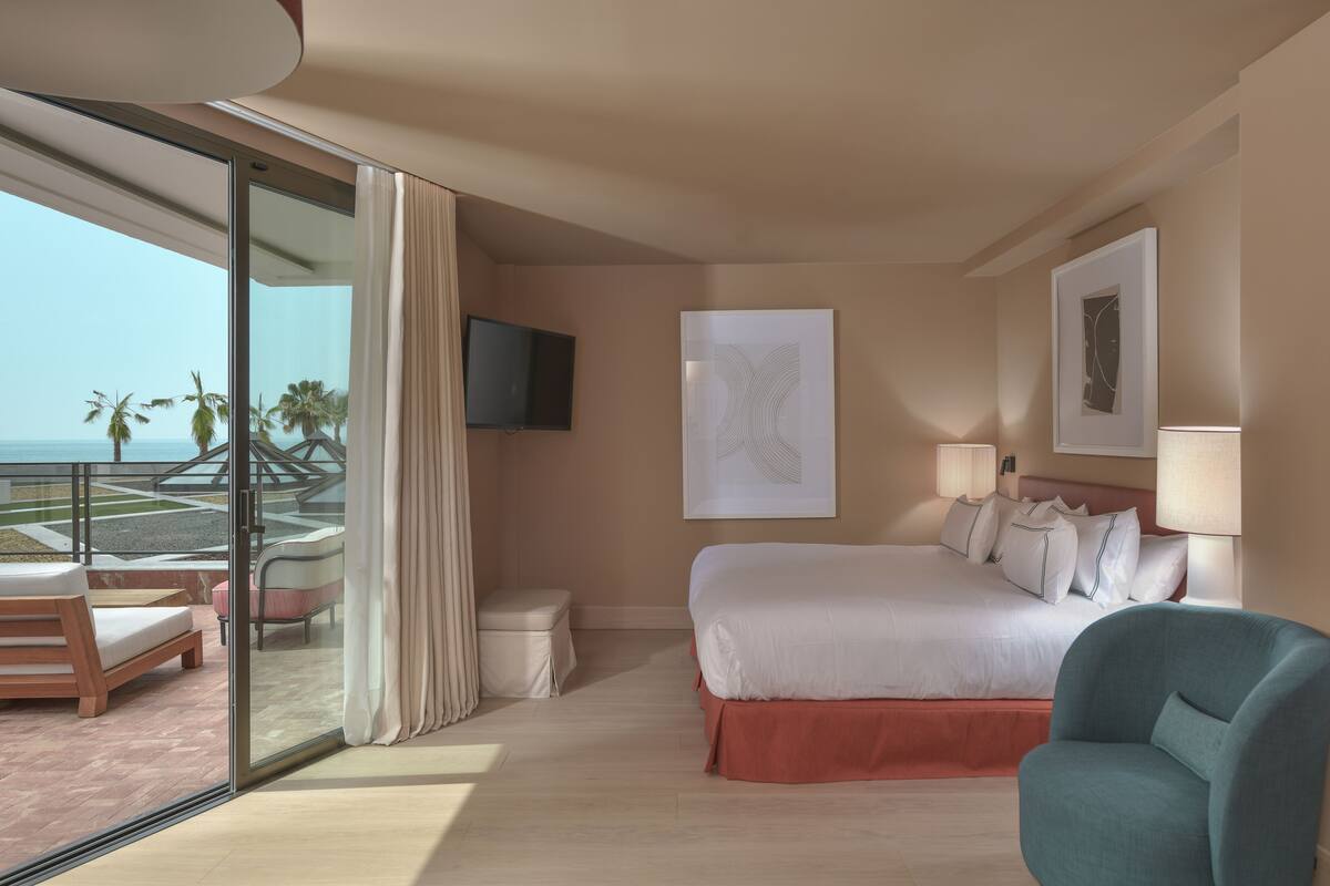 A hotel room with a bed, a chair, a TV, and a view of the beach.