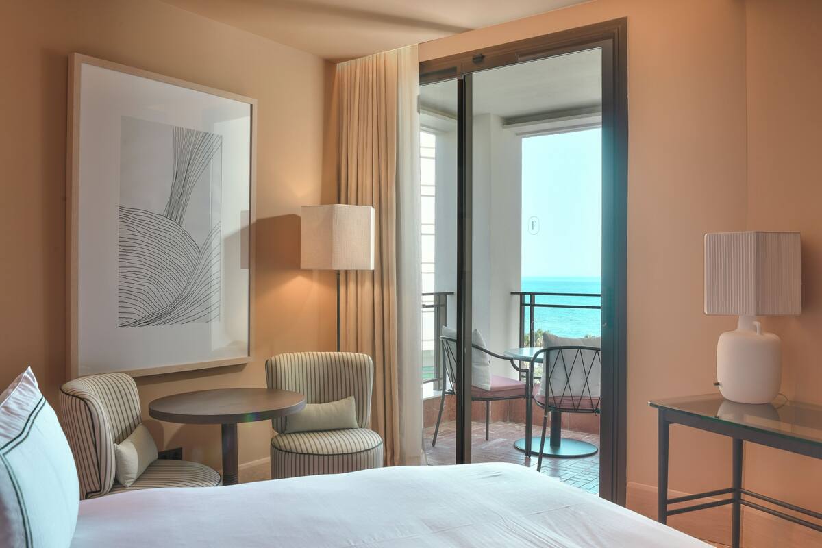 A hotel room with a bed, a desk, a chair, a table, and a balcony with a view of the ocean.