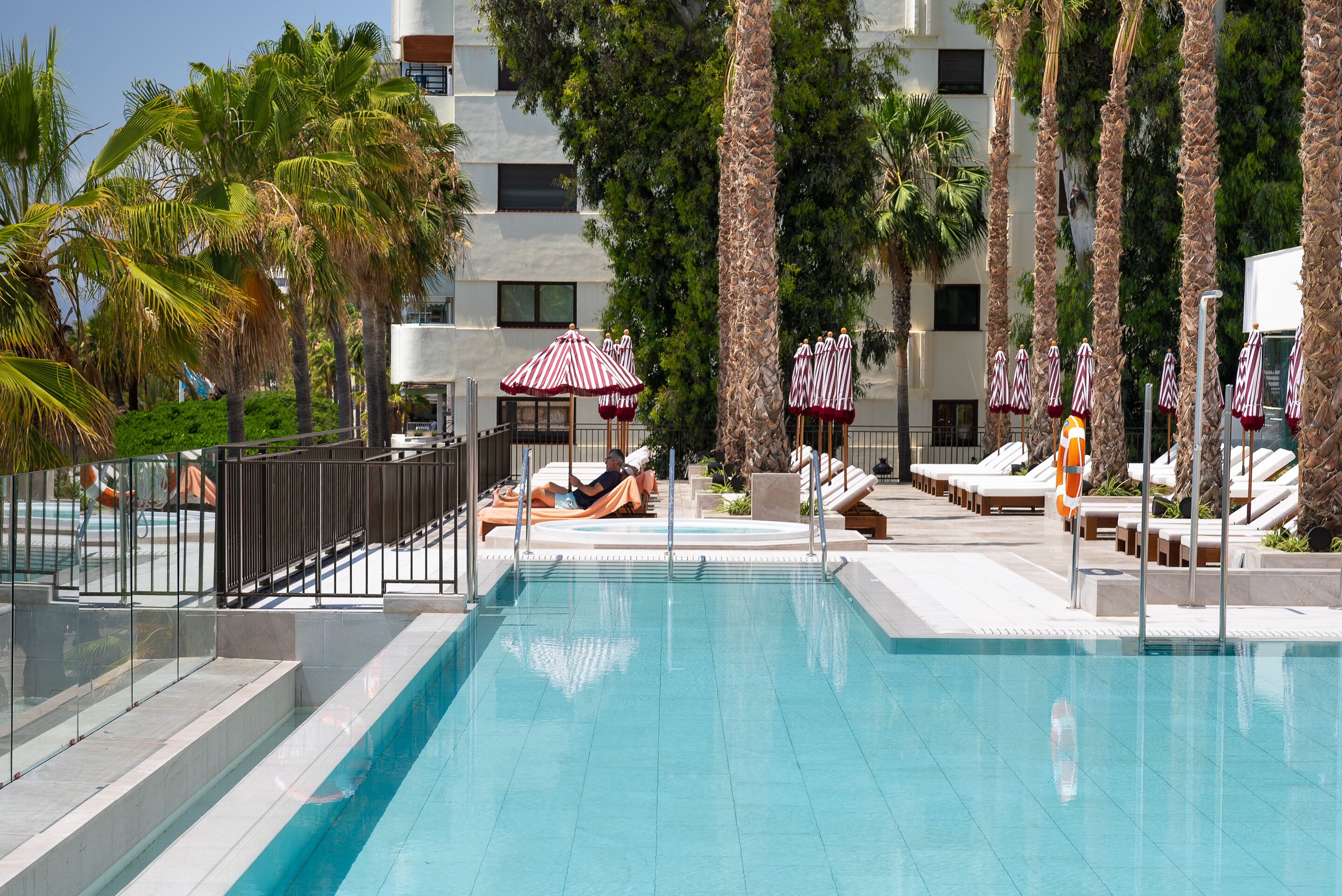 A swimming pool with lounge chairs and umbrellas, surrounded by palm trees and a building with multiple windows.