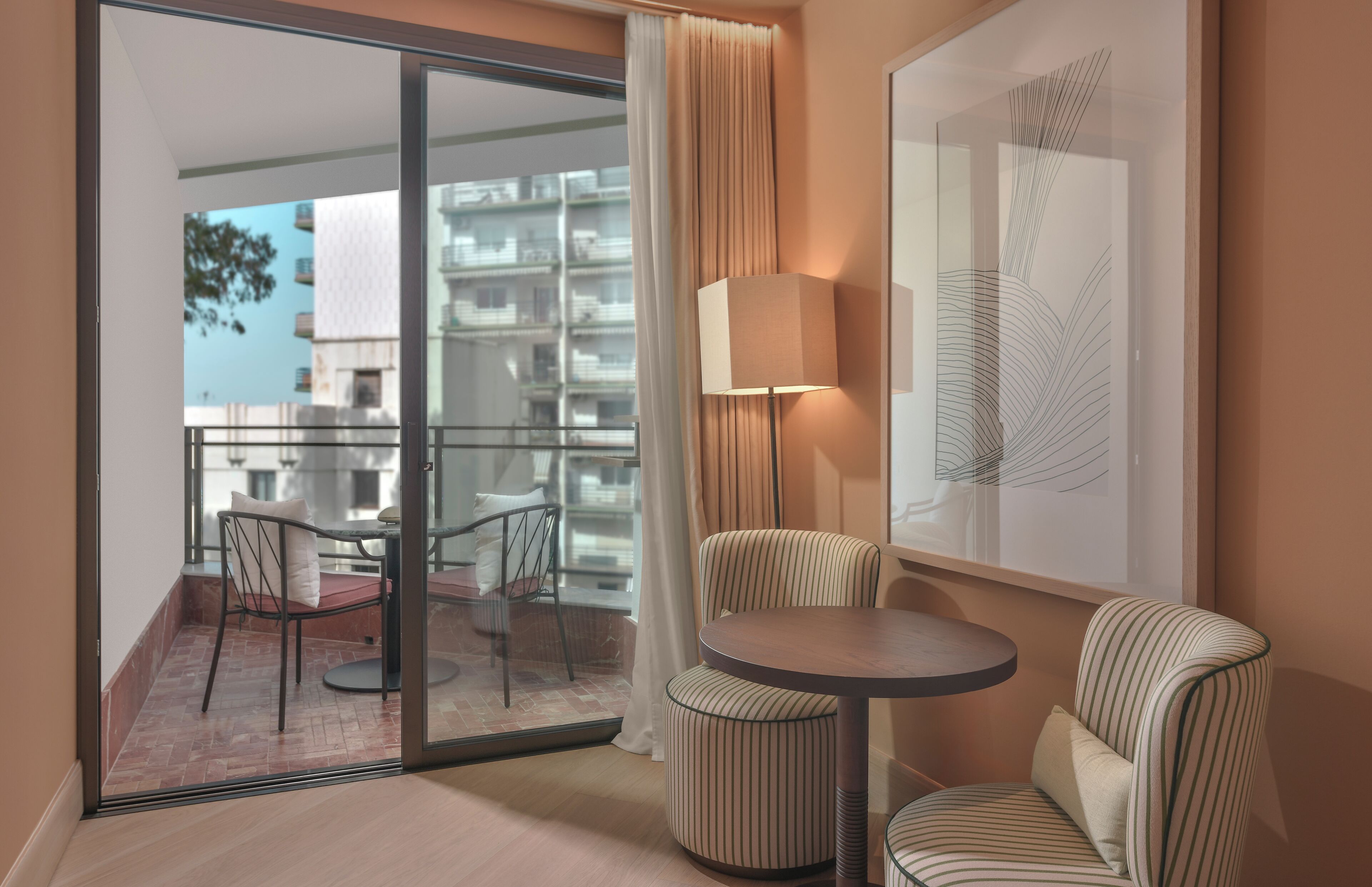 A modern hotel room with a balcony, a round table, two striped chairs, a floor lamp, and a large abstract painting.