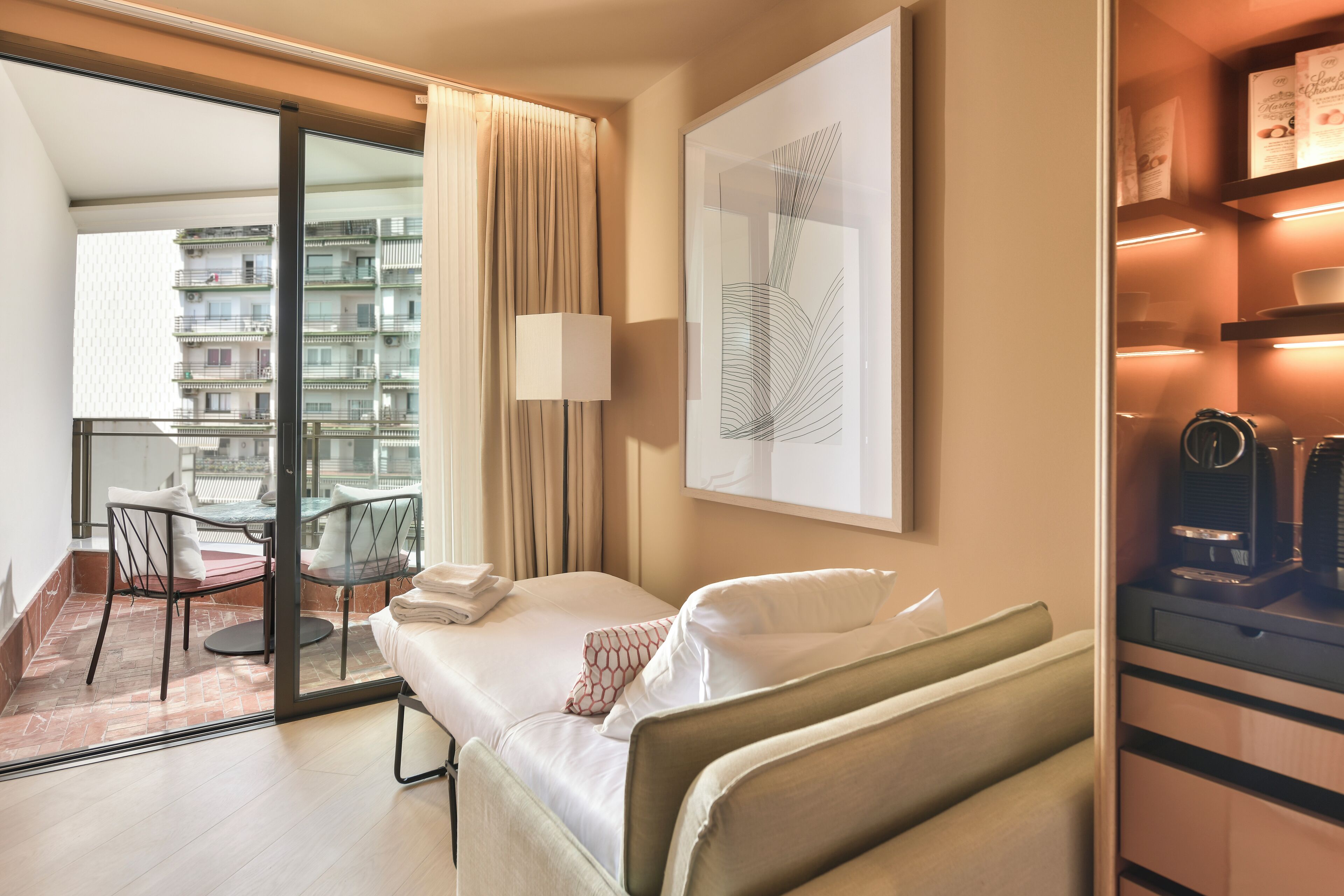 Een hotelkamer met een bed, een bureau, een stoel, een lamp, een raam met gordijnen, een lijst met kunstwerk en een plank met spullen.