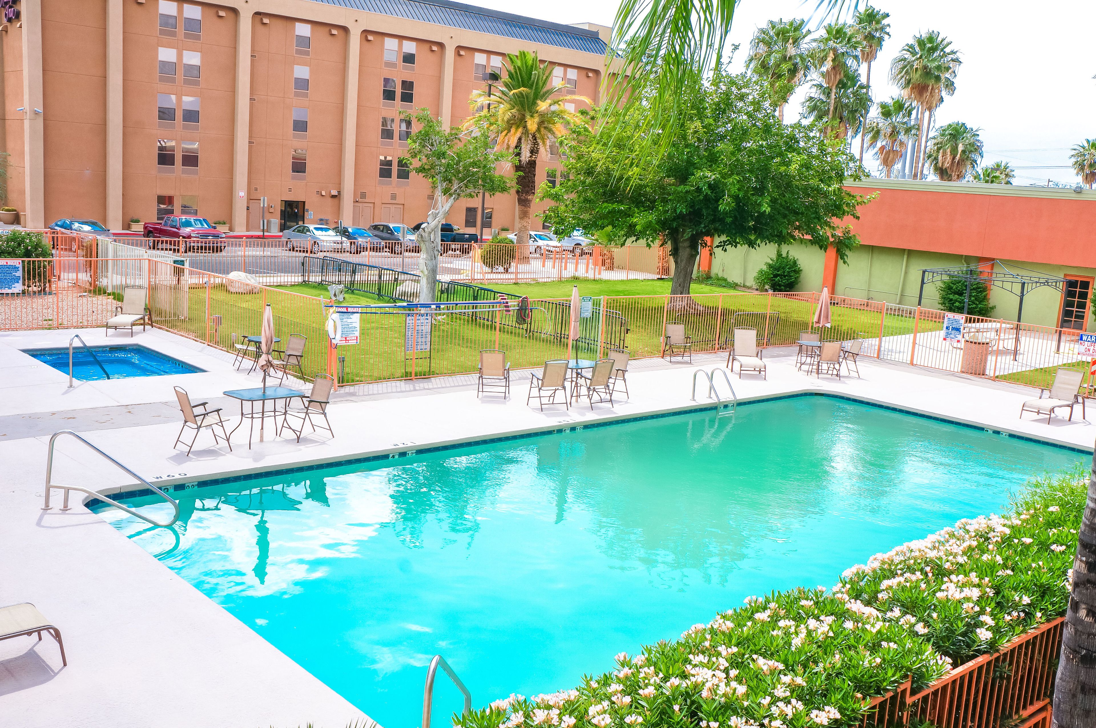 A swimming pool with lounge chairs and a small table, surrounded by a fenced area with palm trees and a building in the background.