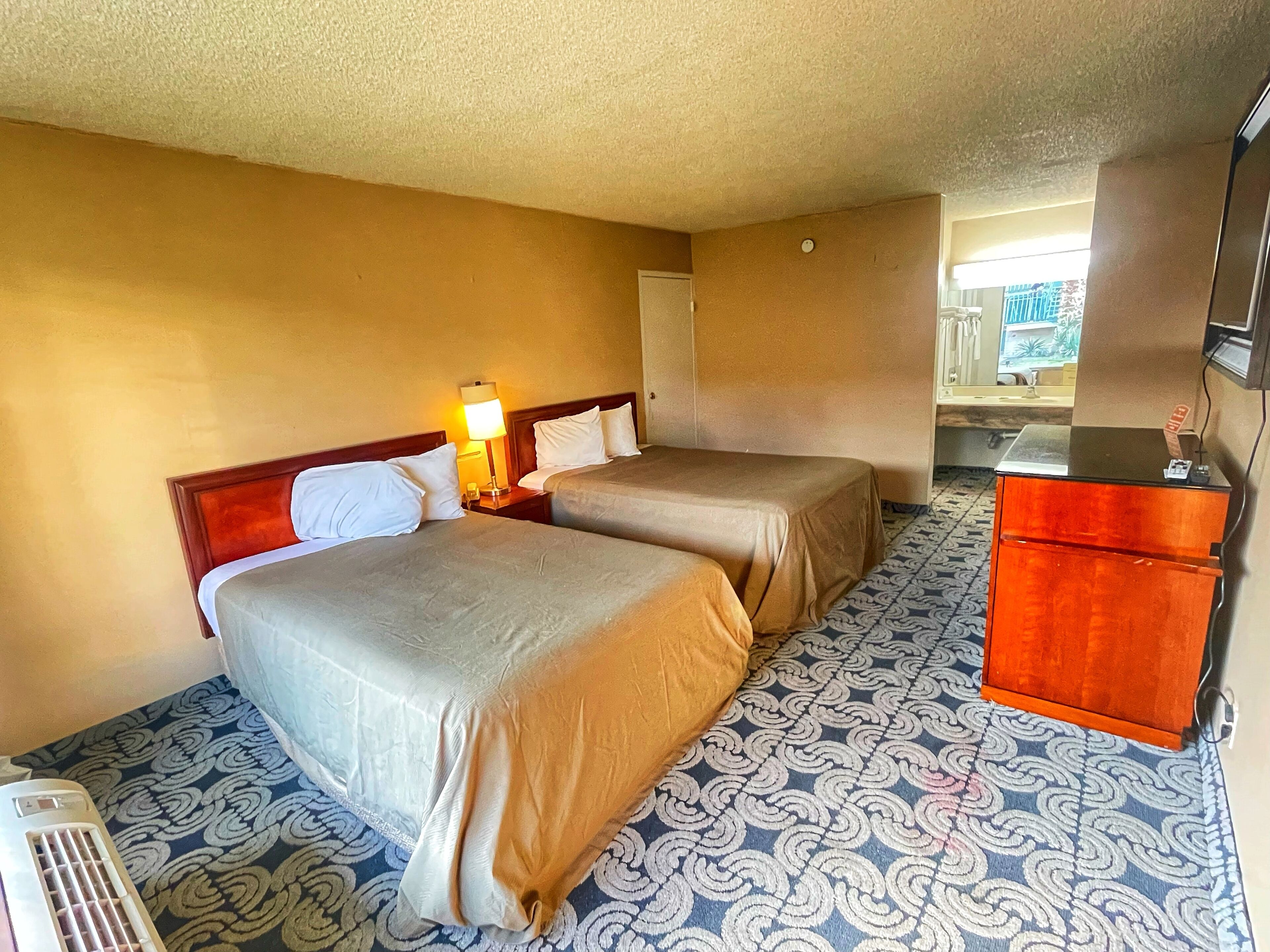 A hotel room with two beds, a bedside table with a lamp, a dresser, and a patterned carpet.