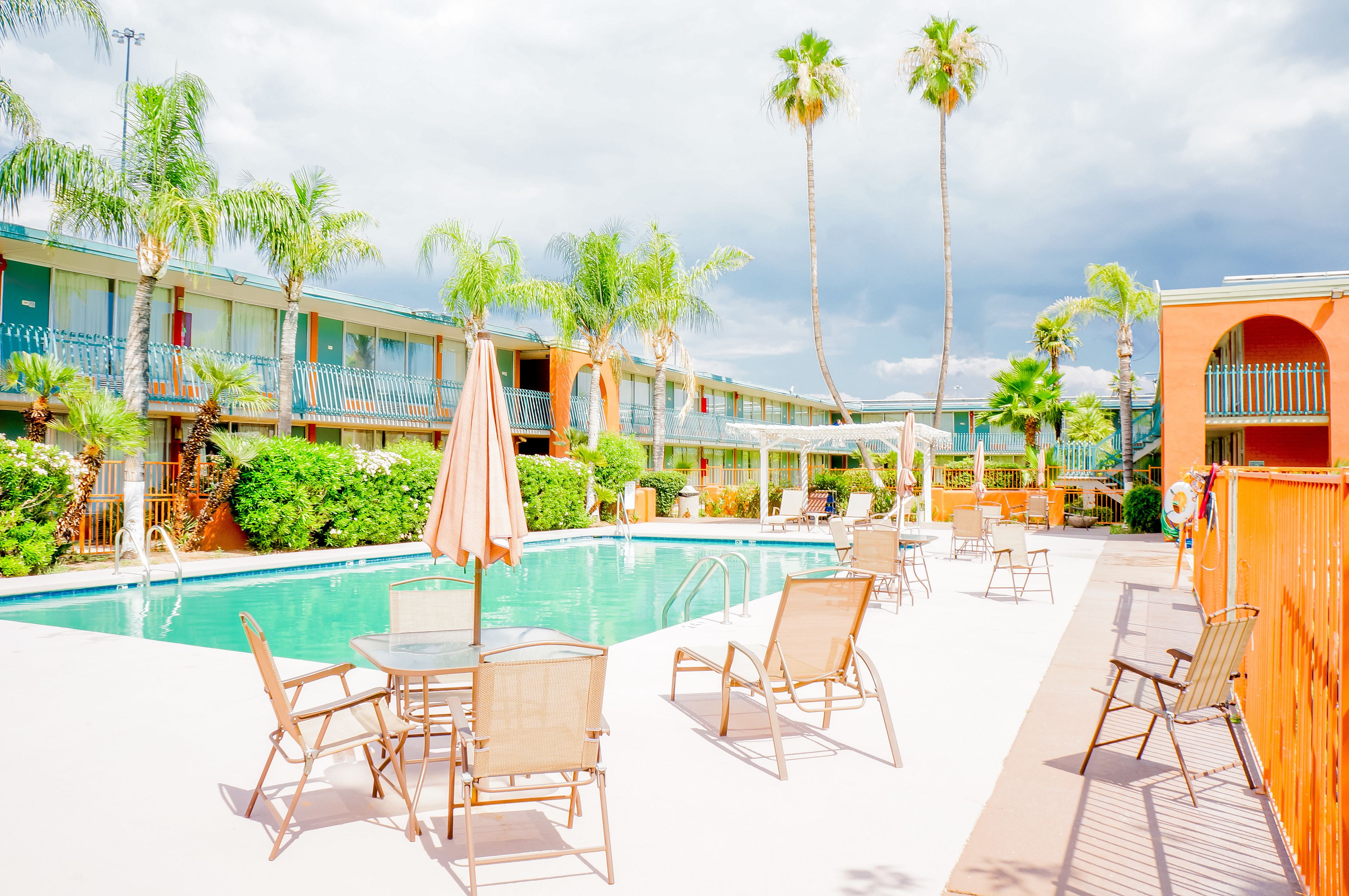Zona de piscina con sillones y sombrillas, rodeada de palmeras y un edificio con balcones.