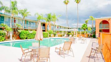 Zona de piscina con sillones y sombrillas, rodeada de palmeras y un edificio con balcones.