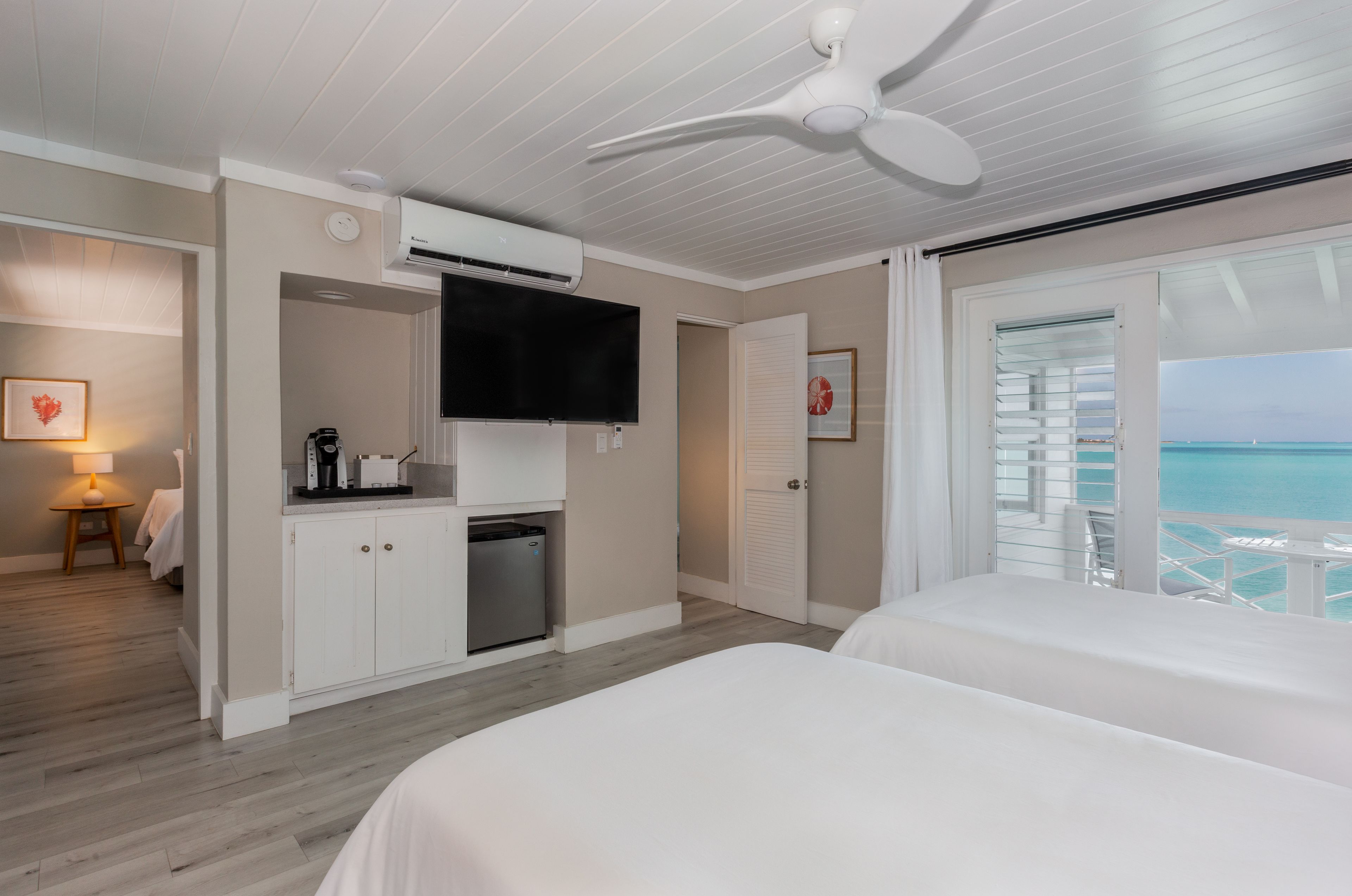 A hotel room with a bed, a TV, a kitchenette, and a balcony with a view of the ocean.