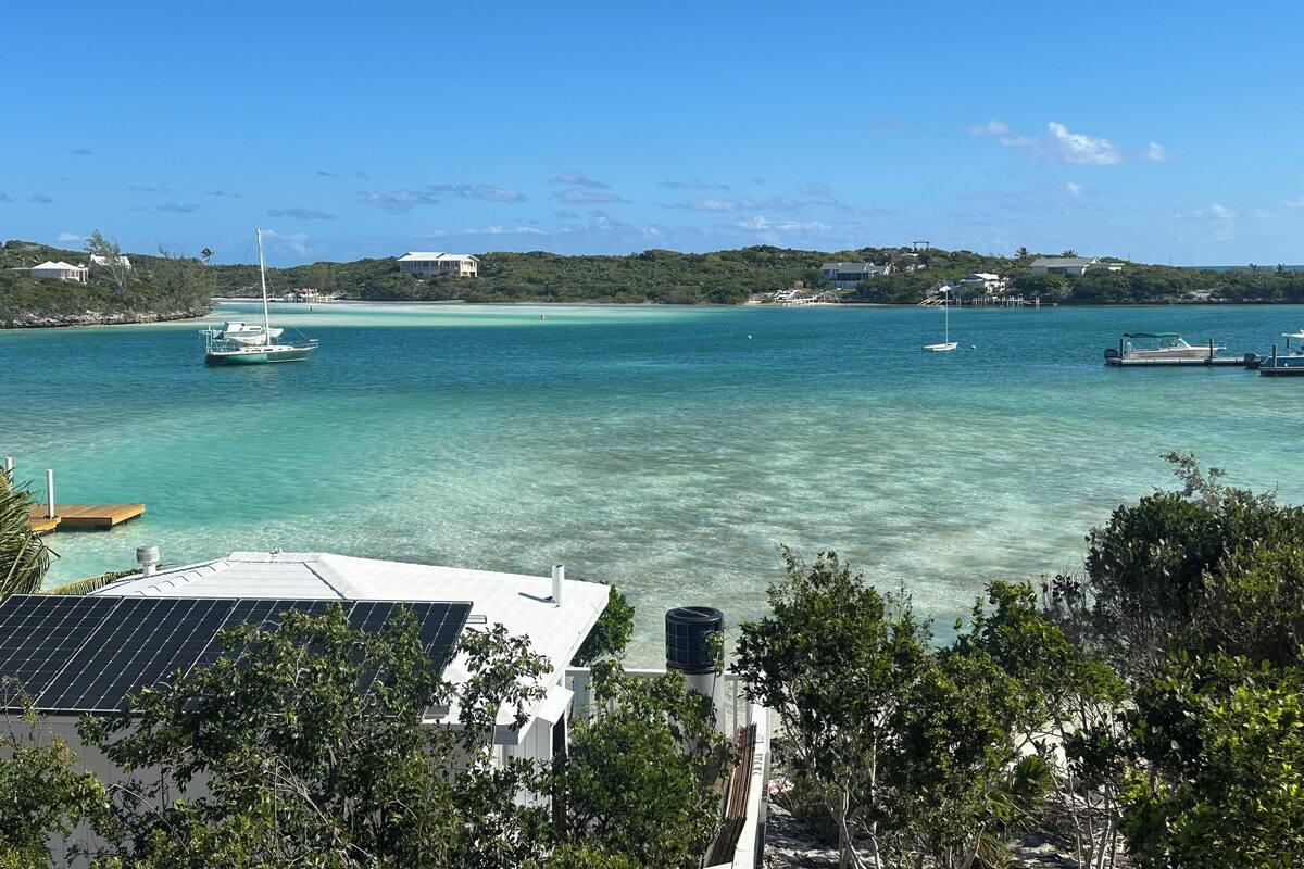 A coastal view with clear turquoise waters, a sailboat, and a house with solar panels.