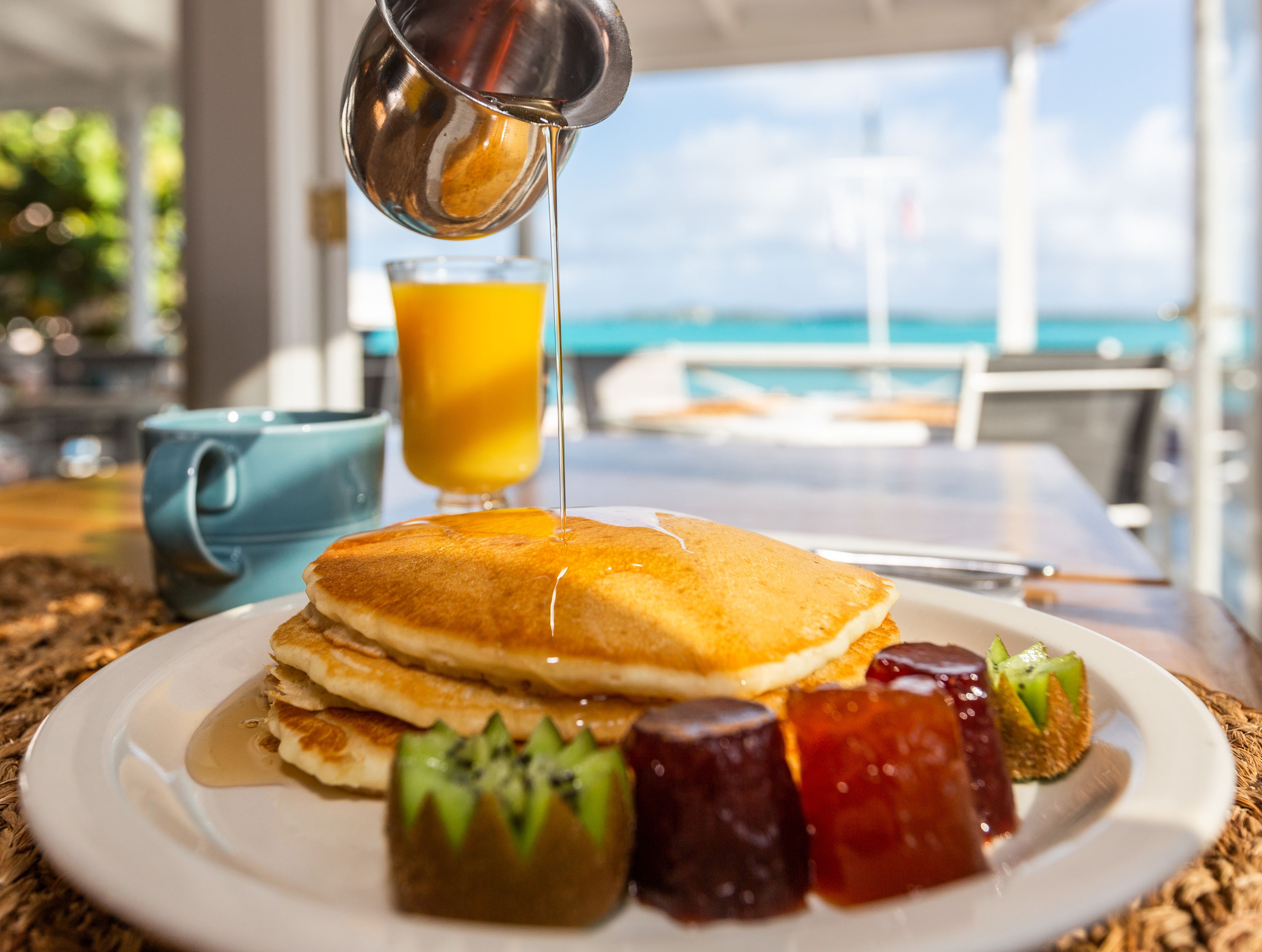 Ein Frühstücksteller mit Pfannkuchen, Sirup und Obst auf einem Tisch mit Meerblick.