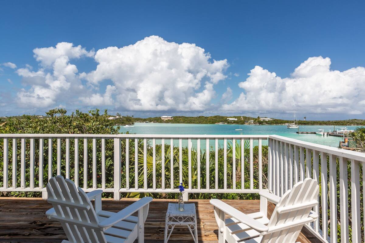 A wooden deck with white railings, two white Adirondack chairs, and a small table with a blue object. The deck overlooks a turquoise sea with boats and a distant shoreline.