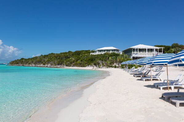 Una playa de arena blanca, agua turquesa cristalina y un edificio blanco con balcón. Hay sombrillas azul y blanco a rayas y sillas de playa blancas.