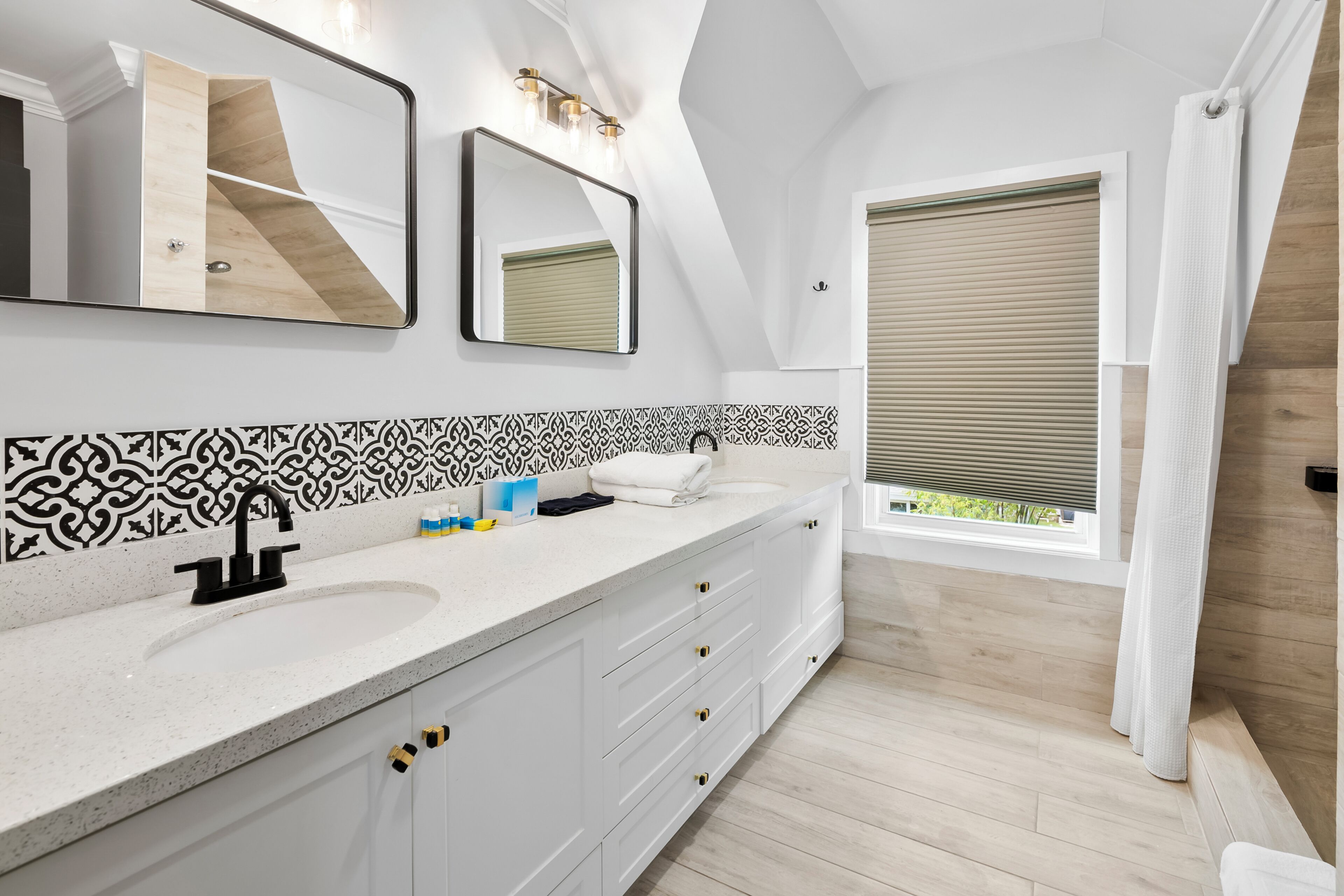 A modern bathroom with a large mirror, two sinks, and a window with blinds.