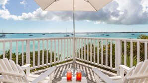 Un balcón con sillas blancas, una mesa con dos vasos y vistas al mar con barcos.