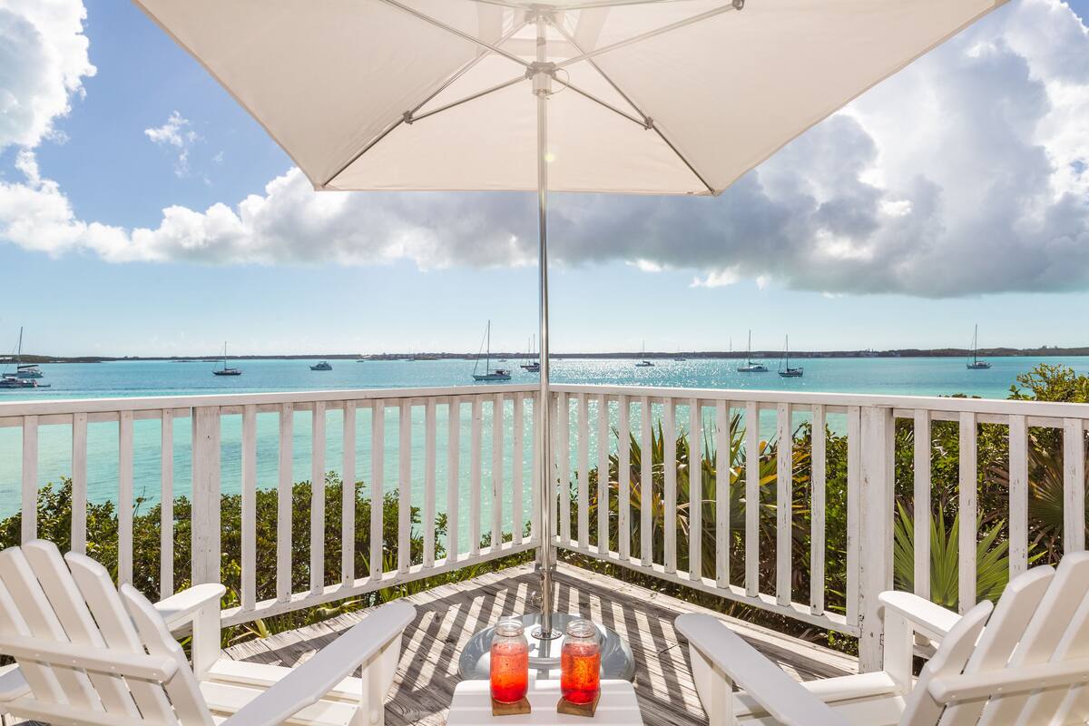 A balcony with white chairs, a table with two glasses, and a view of the sea with boats.