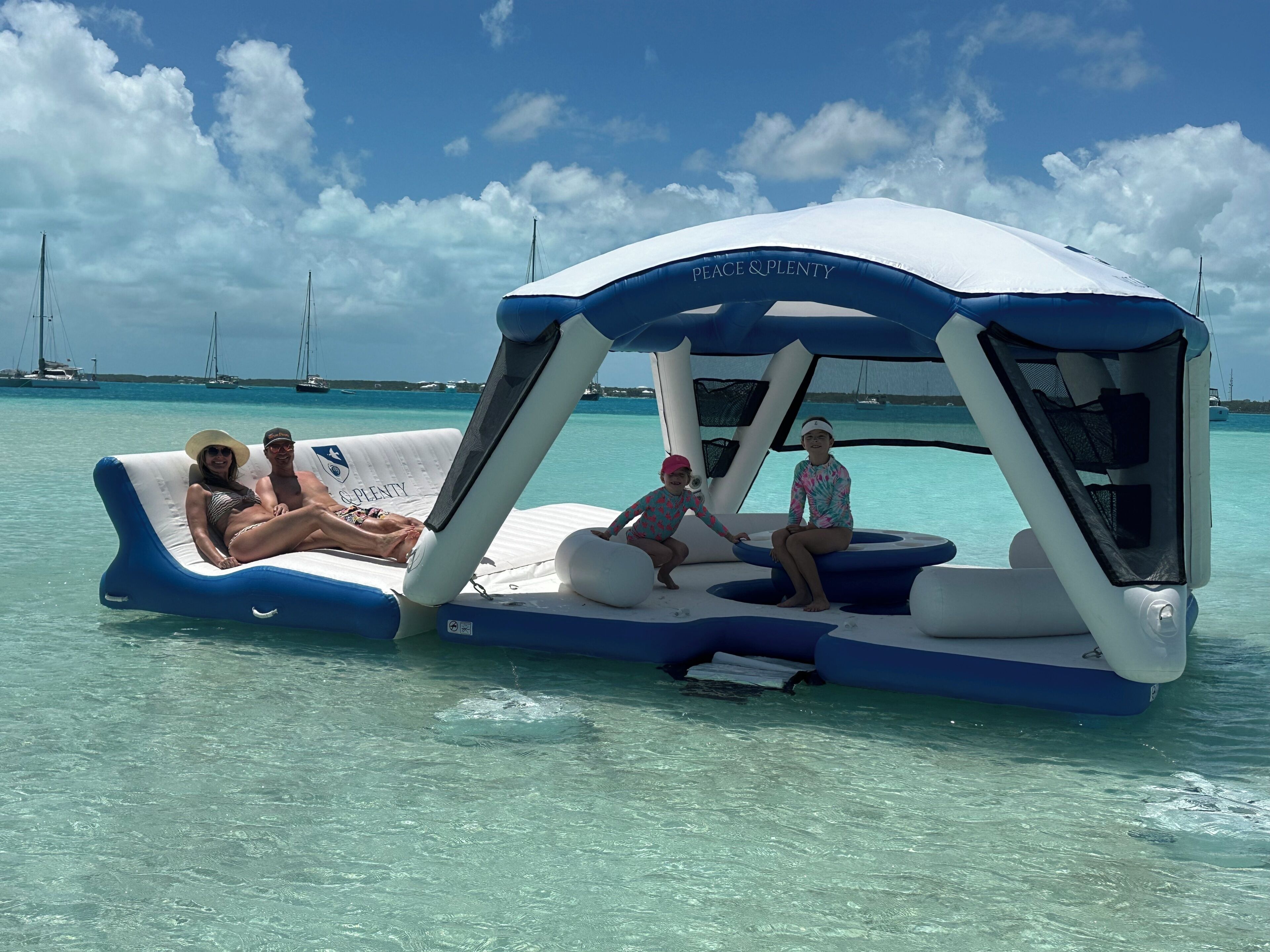 Una plataforma flotante con un toldo que dice 'Peace & Plenty' en un mar azul cristalino, con veleros al fondo.