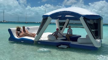 Una plataforma flotante con un toldo que dice 'Peace & Plenty' en un mar azul cristalino, con veleros al fondo.
