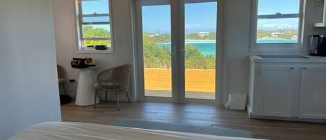A bedroom with a large bed, a chair, a desk, and a view of the sea through a sliding door.