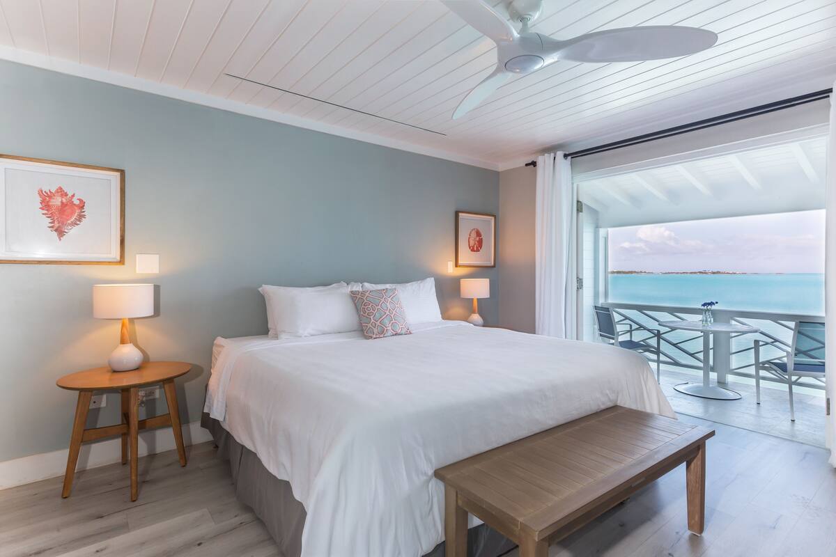 A bedroom with a bed, bedside tables, a bench, and a view of the ocean through a sliding door.