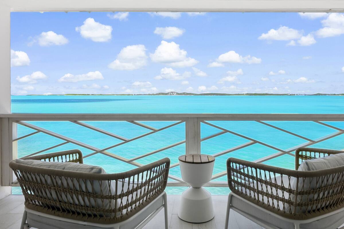 A balcony with wicker chairs and a white table, overlooking a turquoise sea and a clear sky.
