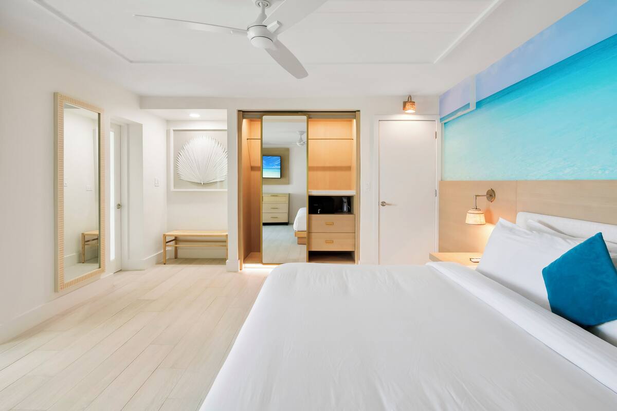 A hotel room with a bed, a ceiling fan, a nightstand, a wall-mounted TV, and a bathroom visible through an open door.