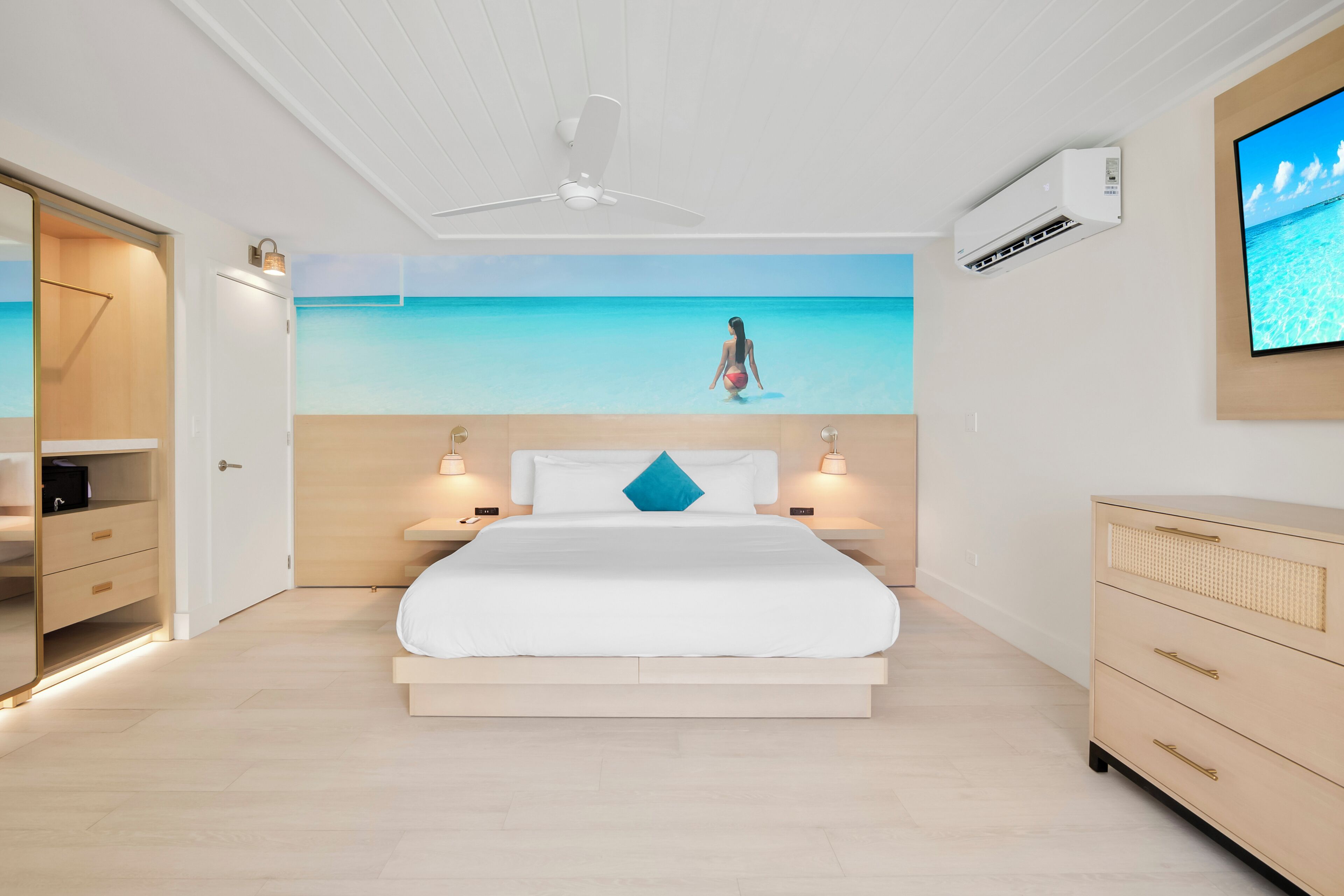 A bedroom with a bed, bedside tables, a TV, and a wall mural of a beach scene.
