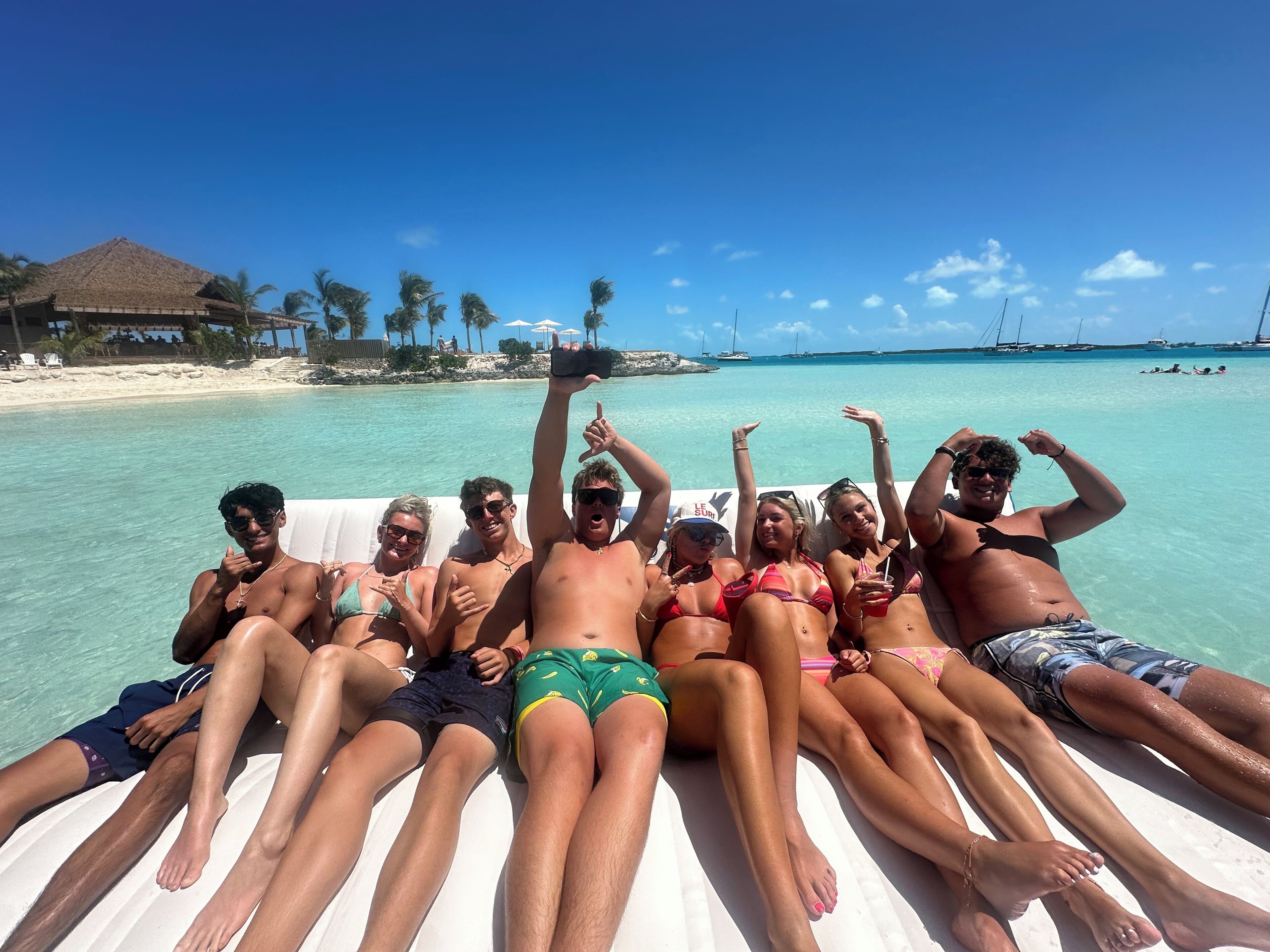 A group of people lounging on a white inflatable raft in a clear turquoise sea, with a beach and palm trees in the background.