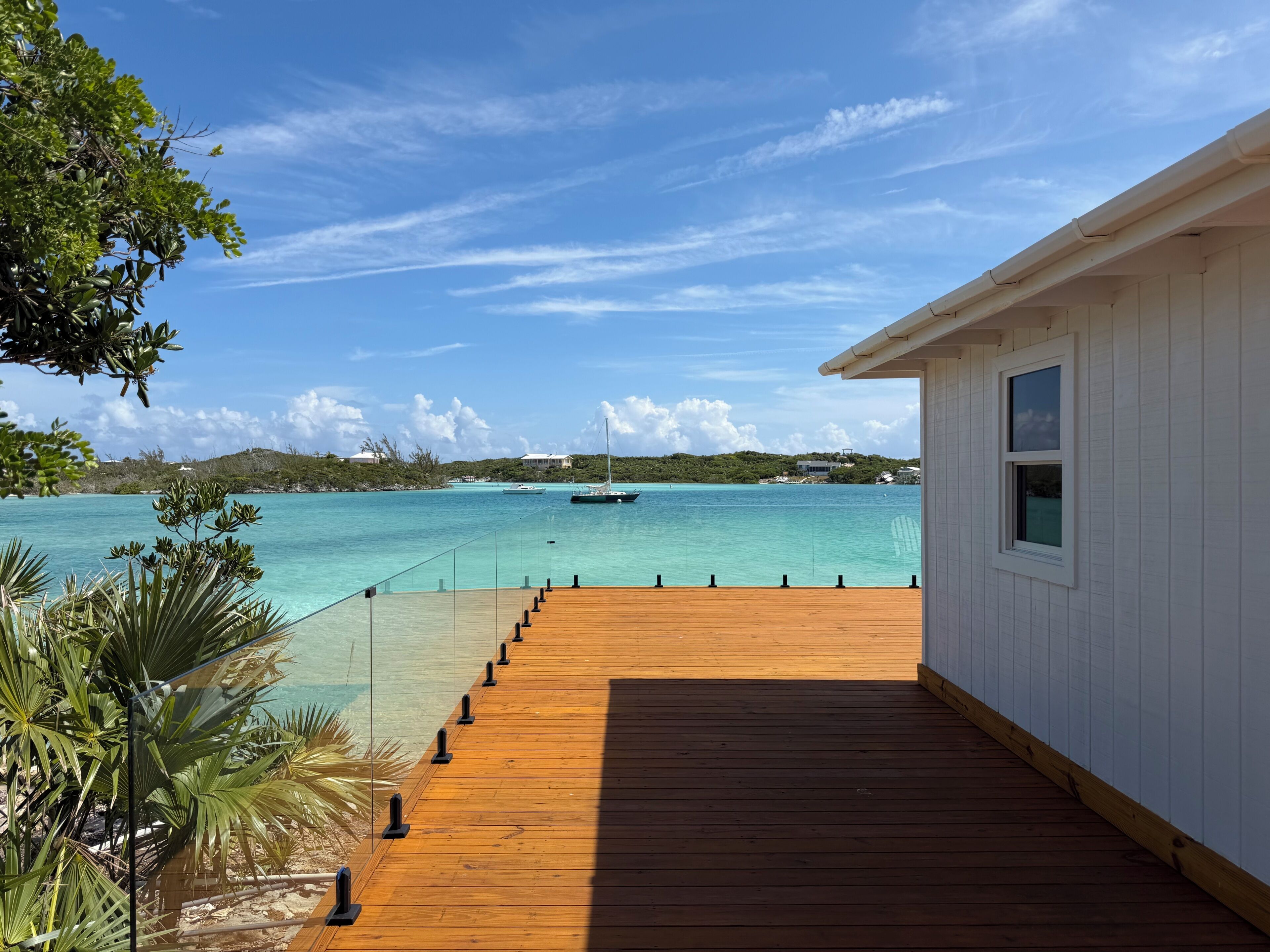 Eine Holzterrasse führt zu einem weißen Gebäude mit Blick auf das türkisfarbene Meer und Boote.