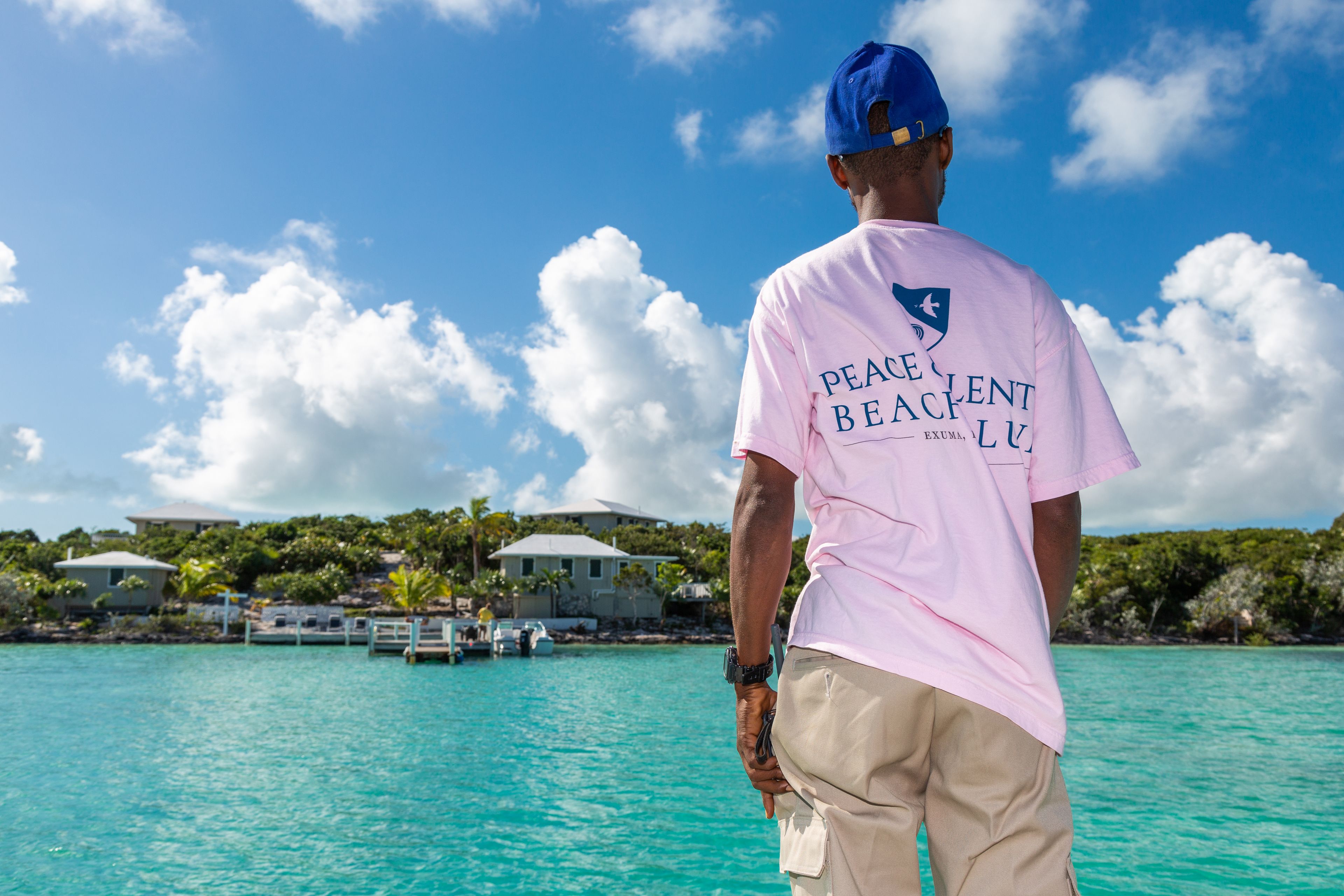 A person standing by a turquoise sea with buildings and trees in the background.
