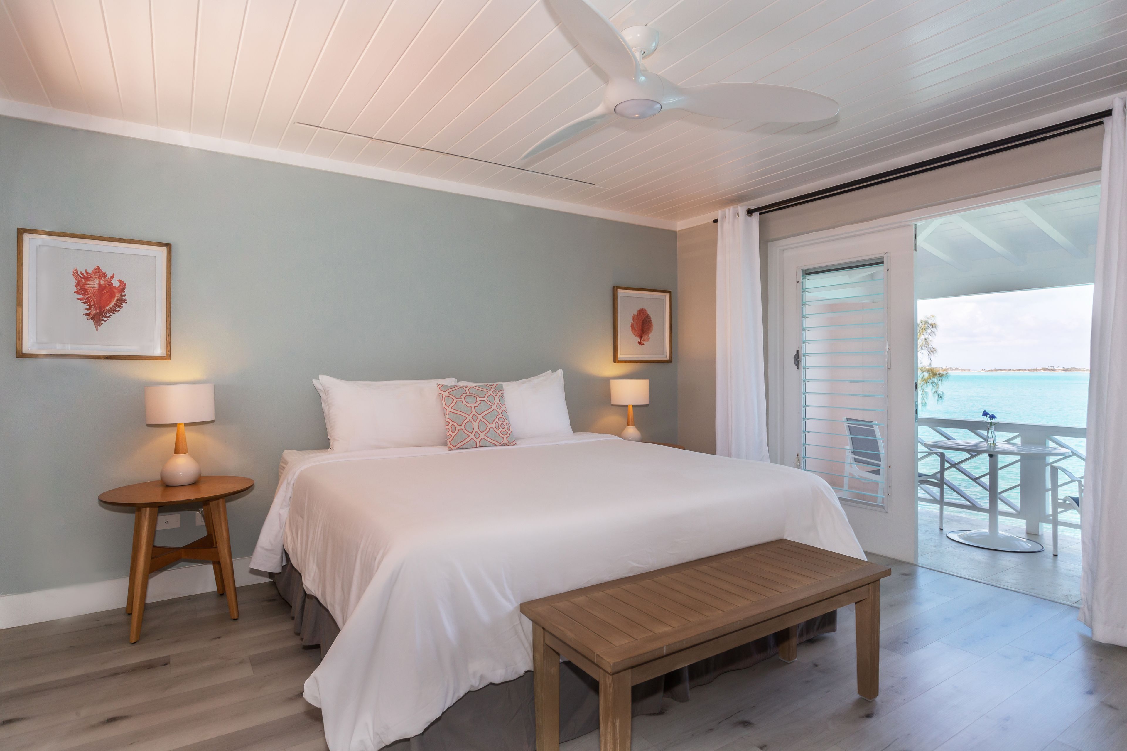 A bedroom with a bed, bedside tables, a bench, ceiling fan, and a balcony with a view.