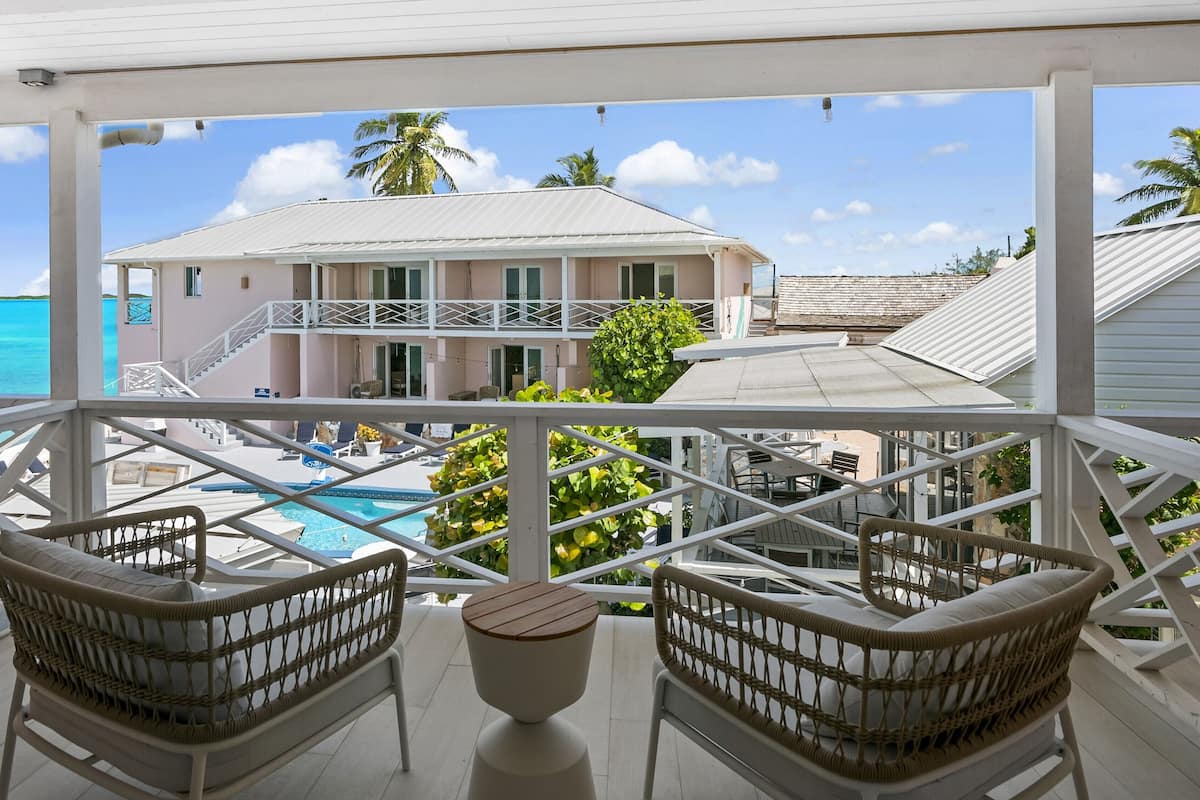 A balcony with wicker chairs and a small table, overlooking a pool and a building with a white facade and a balcony.
