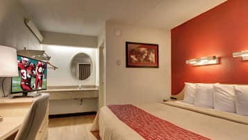A hotel room with a bed, a desk with a computer, a mirror, a framed picture on the wall, and a red accent wall.